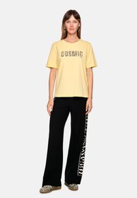 Yellow cotton t-shirt with "COSMIC LOVE" in black, paired with black wide-leg pants featuring a white zebra stripe design on the side.