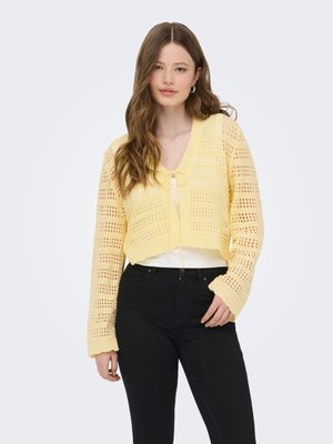 Ung kvinde med langt brunt hår bærer en gul åbenstrikket cardigan over en hvid top og sorte bukser, stående mod en ensfarvet lys baggrund.