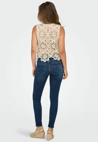 Top de ganchillo crema con diseño de corte en flor, combinado con pantalones vaqueros ajustados oscuros y cuñas de esparto color bronceado. Detalle de abertura en la parte posterior.