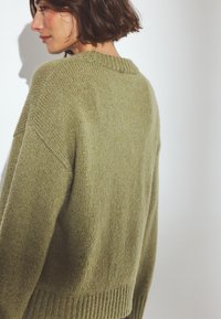 Oliwkowy sweter dzianinowy o luźnym kroju, z prążkowanym okrągłym dekoltem i teksturowanym wykończeniem. Posiada opadające ramiona i szeroki dół.