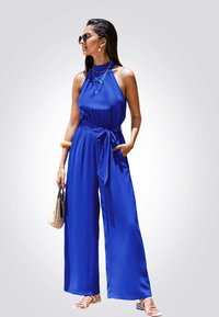 Mono azul sin mangas con cuello halter, cintura ajustada y diseño de pierna ancha. Acentuado con un cinturón y bolsillos, combinado con sandalias y un bolso.