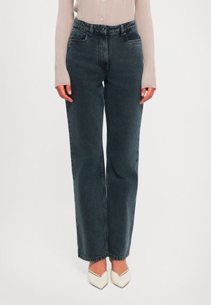 Kvinne iført høytaljede svarte straight-leg jeans, beige ribbet langermet topp og hvite spisse hæler mot hvit bakgrunn.