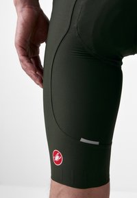 Donkergroene fietsshorts gemaakt van gestructureerde stof, met een strakke pasvorm, zijzak en een rood merkembleem nabij de zoom.