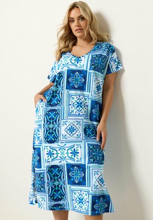 Kobieta ubrana w niebiesko-białą, wzorzystą, długą sukienkę z krótkim rękawem, stoi oparta o białą ścianę, z jedną ręką w kieszeni.