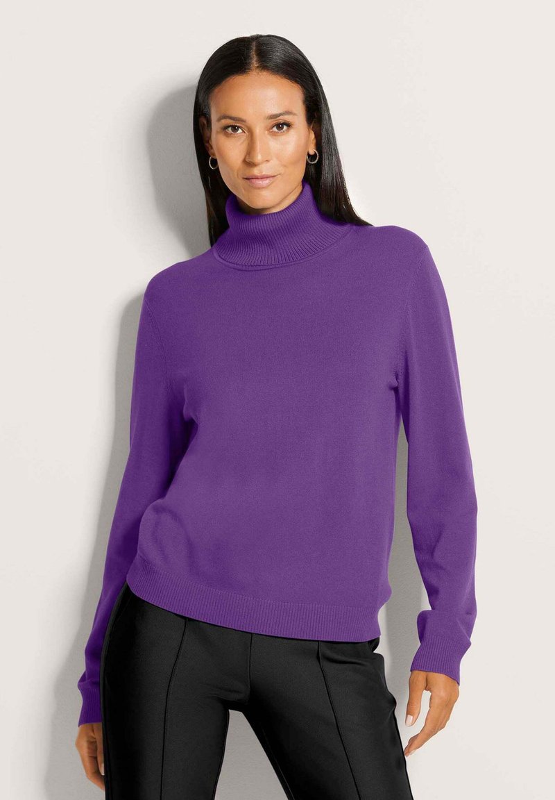 Lila Rollkragenpullover aus weichem Strickmaterial mit gerippten Akzenten am Kragen und Saum. Kombiniert mit schmalen schwarzen Hosen.