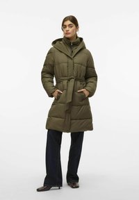 Ženska v olivno zeleni puhovki s pasom in kapuco, temnih širokih hlačah in temnih čevljih s peto, stoji s rokami v žepih.