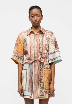 Γυναίκα με φόρεμα πουκάμισο με σχέδια και ζώνη, φαρδιά μανίκια σε γήινους τόνους και παστέλ μπλε, στέκεται μπροστά σε απλό λευκό φόντο.