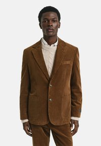 Blazer en velours côtelé marron avec deux poches avant et une poche sur la poitrine. Porté par-dessus un pull en maille torsadée crème et une chemise blanche.