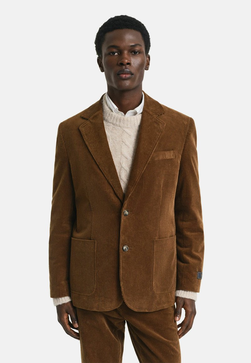 Blazer en velours côtelé marron avec deux poches avant et une poche sur la poitrine. Porté par-dessus un pull en maille torsadée crème et une chemise blanche.