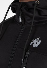 Zwarte zip-up jack gemaakt van soepel materiaal, met een zilver gorilla-logo en een ritszak. Inclusief verstelbare trekkoorden bij de capuchon.