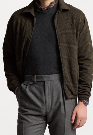 Homme portant une veste marron à carreaux zippée sur un pull noir et un pantalon gris plissé, main dans la poche et portant une montre-bracelet.