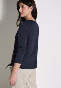 Maglietta a maniche lunghe blu navy con scollatura rotonda, tessuto morbido e un accento annodato in vita, abbinata a pantaloni beige chiaro.