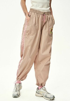 Osoba ubrana w beżowe szerokie spodnie dresowe z różowymi bocznymi panelami, elastycznymi mankietami i różowymi sznurowadłami, w połączeniu ze srebrnymi trampkami i różowymi skarpetkami.