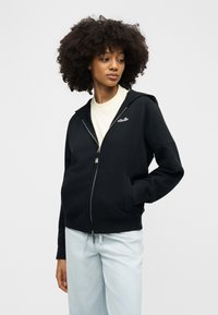 Γυναίκα με σγουρά μαλλιά που φοράει μαύρο φούτερ με φερμουάρ πάνω από λευκό πουκάμισο και ανοιχτό γαλάζιο παντελόνι, στάζει με το ένα χέρι στην τσέπη και κοιτάζει πλάγια.