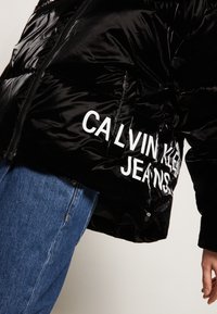 Černá péřová bunda s lesklým povrchem, se značkovým nápisem "CALVIN KLEIN JEANS" v bílé barvě a zipovým zapínáním. V kombinaci s modrými džíny.