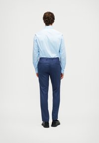 Άνδρας στέκεται με την πλάτη στραμμένη, φορώντας ανοιχτό μπλε μακρυμάνικη πουκάμισο, σκούρα μπλε παντελόνια προσαρμοσμένα και μαύρες κοσμικές παπούτσια σε απλό φόντο.