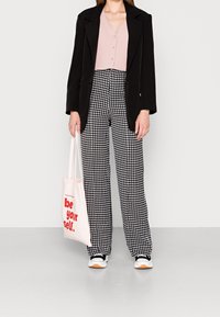 Blazer noir sur un haut rose clair à boutons, associé à un pantalon large à motif pied-de-poule noir et blanc, et portant un sac fourre-tout rose avec du texte rouge.