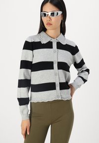 Siv in črn cardigan s črtami, s čipkastim ovratnikom, sprednjimi gumbi in rebrastimi manšetami, v kombinaciji z olivno zelenimi hlačami.