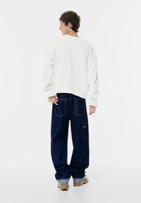 Weißer, gerippter Pullover mit langen Ärmeln, kombiniert mit lockeren dunkelblauen Jeans mit Gesäßtaschen und Nähdetails.