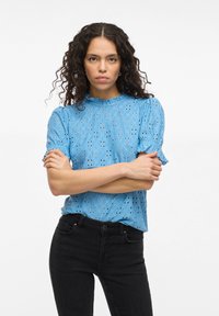 Lyseblå, kortermet topp med volanger i halsen og blondermønster; brukt med svarte høye jeans.