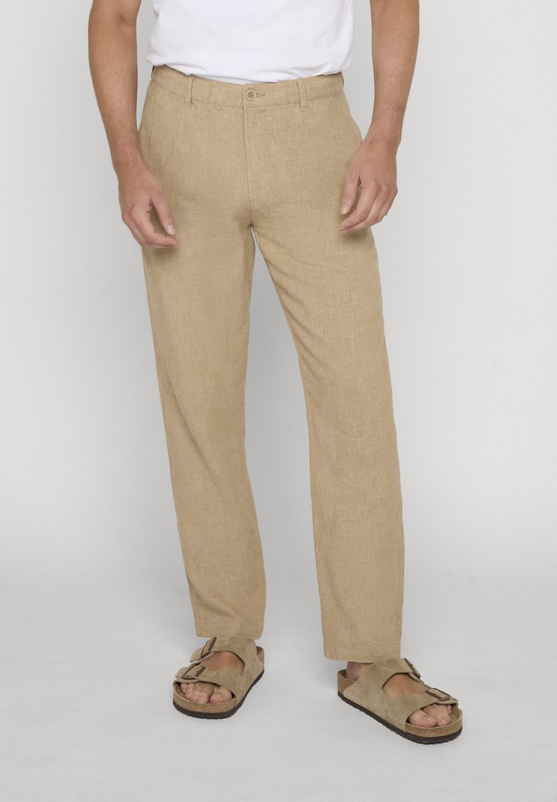 Homme portant un pantalon beige à jambes droites, une chemise blanche et des sandales beige à boucle, debout devant un fond clair uni.