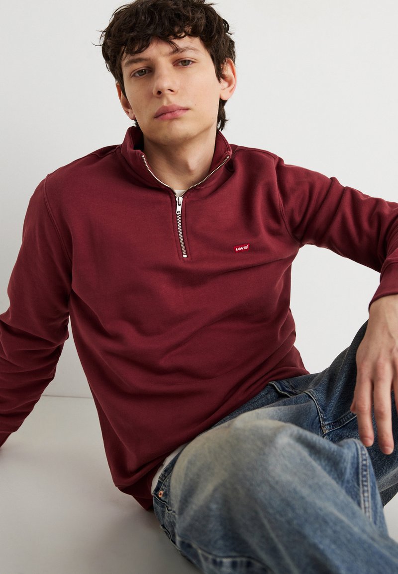 Νεαρός άνδρας που φοράει φούτερ με φερμουάρ μέχρι το στήθος σε μπορντό χρώμα και μπλε τζιν, καθισμένος μπροστά σε απλό ανοιχτόχρωμο φόντο, κοιτάζοντας ελαφρώς προς τα πάνω.