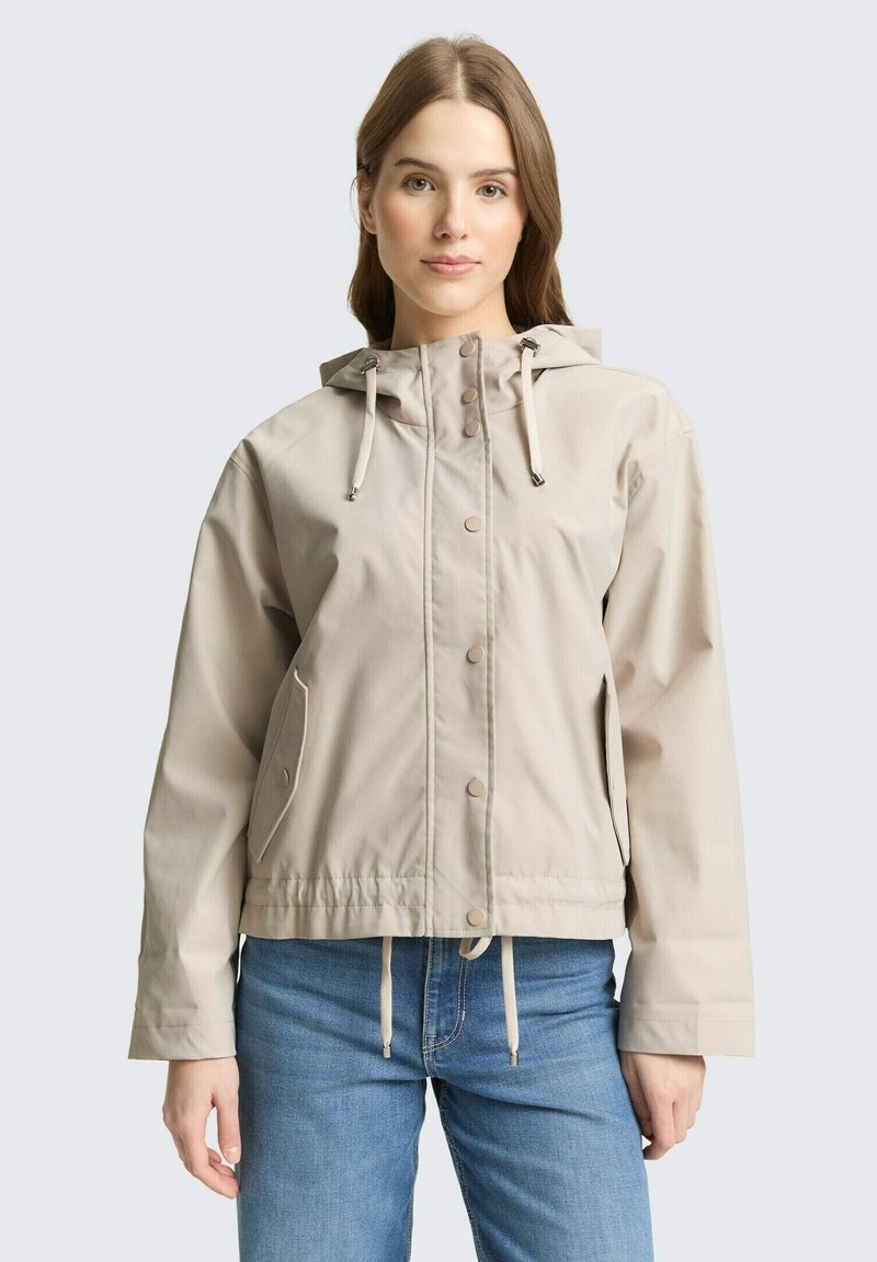 Mladá žena v béžovej kapucňovej bunde so zapínaním na patentky a modrých džínsoch, stojaca pred jednoduchým svetlým pozadím.