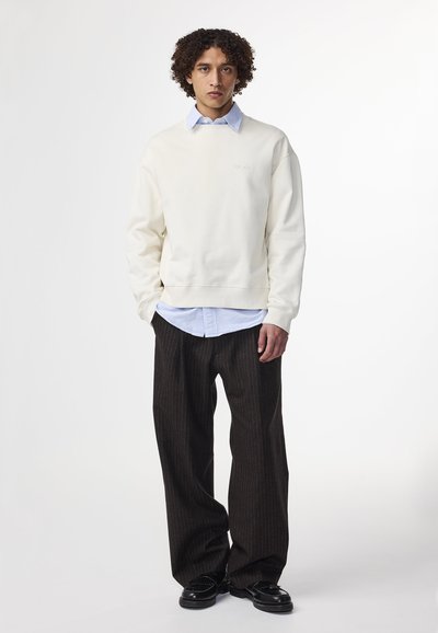 Helles beige Sweatshirt mit gerippten Bündchen, über ein blau-weiß gestreiftes Hemd geschichtet. Das Model trägt dunkel gestreifte Hose und schwarze Schuhe.