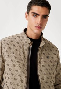 Veste bomber beige à motifs avec un design en grille de losanges, dotée d'une fermeture éclair à l'avant, d'accents marron et d'un logo sur la poitrine.