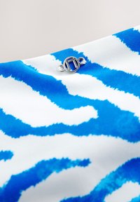 Gros plan sur un tissu rayé bleu et blanc avec un petit emblème métallique argenté à motif circulaire cousu dessus.