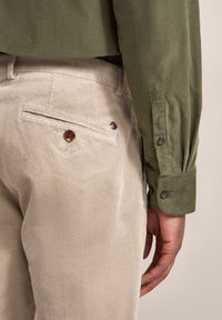Khaki Cord-Leinen-Hosen mit einer Gesäßtasche, ausgestattet mit einem dekorativen Knopf mit roten und blauen Akzenten. Olivgrünes Langarmhemd aus strukturiertem Stoff.