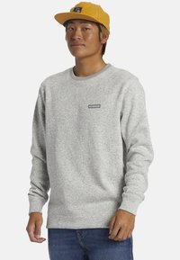 Grauer Sweatshirt mit strukturiertem Finish und geripptem Rundhals-Ausschnitt. Verfügt über ein kleines Logo-Patch auf der Brust. Getragen mit blauen Jeans und einer gelben Mütze.