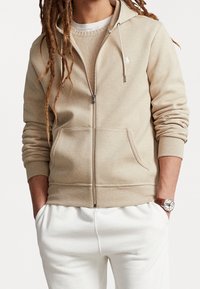Beige zip-up hoodie made of soft fabric, featuring a front pocket and a small white logo on the left chest. Paired with white pants.