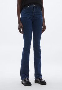 Jeans evazați din denim albastru închis, cu talie înaltă, având un design clasic cu cinci buzunare și o textură netedă. Purtați cu pantofi slip-on negri.