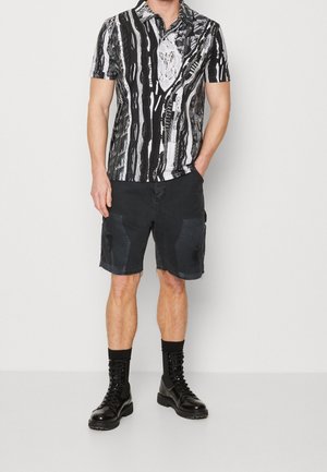 Mann trägt ein schwarz-weiß gestreiftes Kurzarm-Poloshirt, schwarze, abgenutzte Shorts, schwarze Socken und glänzende schwarze Schnürstiefel, steht an einer weißen Wand.