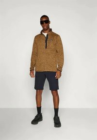 Pruun fliistezippingiga jakker kõrge kaelusega, koos tumesinine lühikeste pükstega ja mustade spordijalatsitega. Mudel kannab päikeseprille, mustreid pole nähtaval.