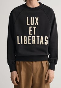 Felpa nera con polsini e orlo a coste, caratterizzata da un grande testo beige che recita "LUX ET LIBERTAS" sul davanti. Tessuto liscio, design con collo a giro.