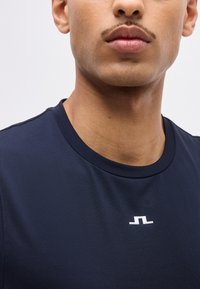 Κοντινό πλάνο ενός ατόμου που φοράει μπλε σκούρο μπλουζάκι με στρογγυλή λαιμόκοψη και ένα μικρό λευκό γεωμετρικό λογότυπο στο κέντρο του στήθους.