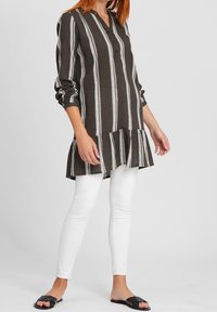 Pasiasta tunika w ciemnym brązie z jasnoszarymi liniami, z guzikowym dekoltem i falbanistym dołem. Połączona z białymi leginsami i czarnymi klapkami.