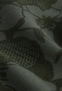 Gros plan sur un tissu tricoté vert foncé avec des motifs floraux texturés et des styles de tissage variés créant des zones en relief et lisses.