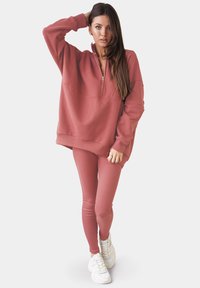Rosafärgad sweatshirt med halv dragkedja och ribbade ärmslut, tillsammans med matchande strukturerade leggings och vita sneakers.
