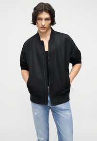 Νέος με μεσαίου μήκους σκούρα μαλλιά φοράει μαύρο bomber μπουφάν πάνω από μαύρο τοπ και ανοιχτό μπλε σκισμένο τζιν, με τα χέρια στις τσέπες.