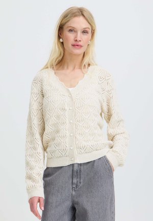 Femme blonde porte un cardigan en tricot crème boutonné et un jean gris taille haute, debout avec une main dans la poche sur un fond uni.