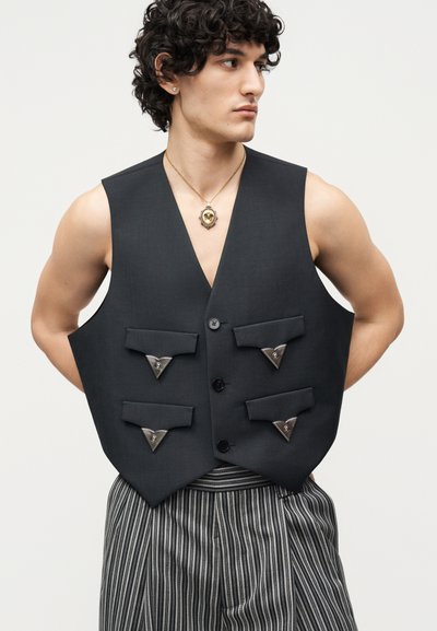 Jeune homme aux cheveux bouclés portant un gilet noir avec quatre poches à rabat et des accents métalliques triangulaires, associé à un pantalon gris rayé.