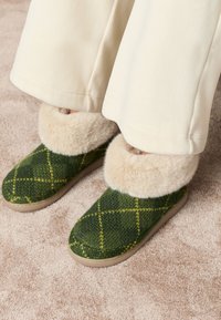 Grønne uldne hjemmesko med diamantmønster, med en fluffy beige falsk pelskant. Blød sål og hyggelig tekstur, placeret på et beige tæppe.