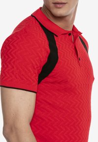 Rotes strukturiertes Polohemd mit einem Zickzackmuster und schwarzen Akzenten an den Schultern. Klassischer Kragen mit zwei Knöpfen am Hals.