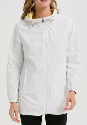 Femme portant une veste à capuche blanche zippée avec doublure jaune et poignets élastiques, debout devant un fond uni.