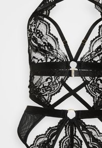 Bralette de encaje negro con intrincados patrones florales, tirantes elásticos, un detalle circular de hardware dorado y diseño de espalda abierta.