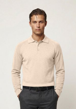 Giovane uomo con capelli pettinati all'indietro che indossa una polo beige a maniche lunghe e pantaloni grigio scuro, in piedi con le mani nelle tasche.