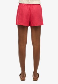 Shorts in lino rosa con vita elastica, visti da dietro. Vita piatta e lunghezza corta, abbinati a sandali marroni.